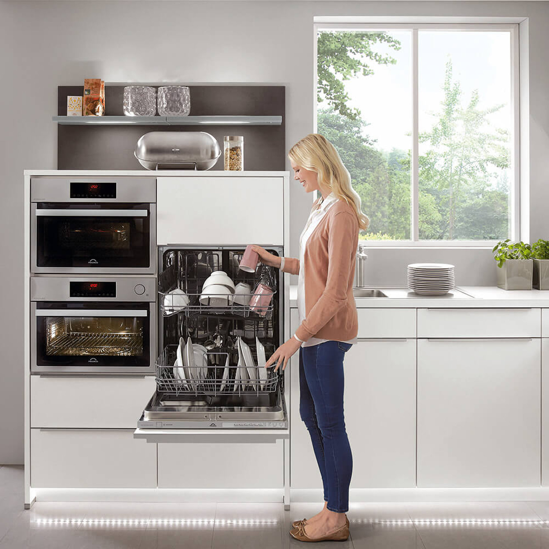 Geräuscharme Küchengeräte: Frau räumt einen höher eingebauten Geschirrspüler in heller, moderner Küche mit Fenster ein.
