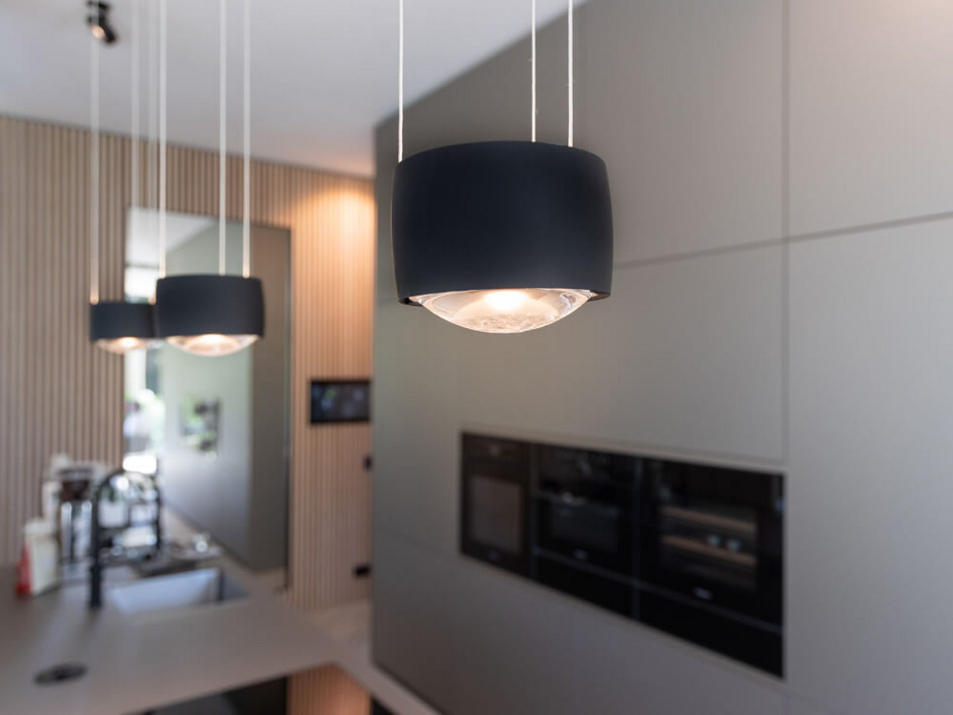 Drei eingeschaltete schwarze Pendelleuchten vor einer grauen Küchenfront und einer Holzlamellenwand