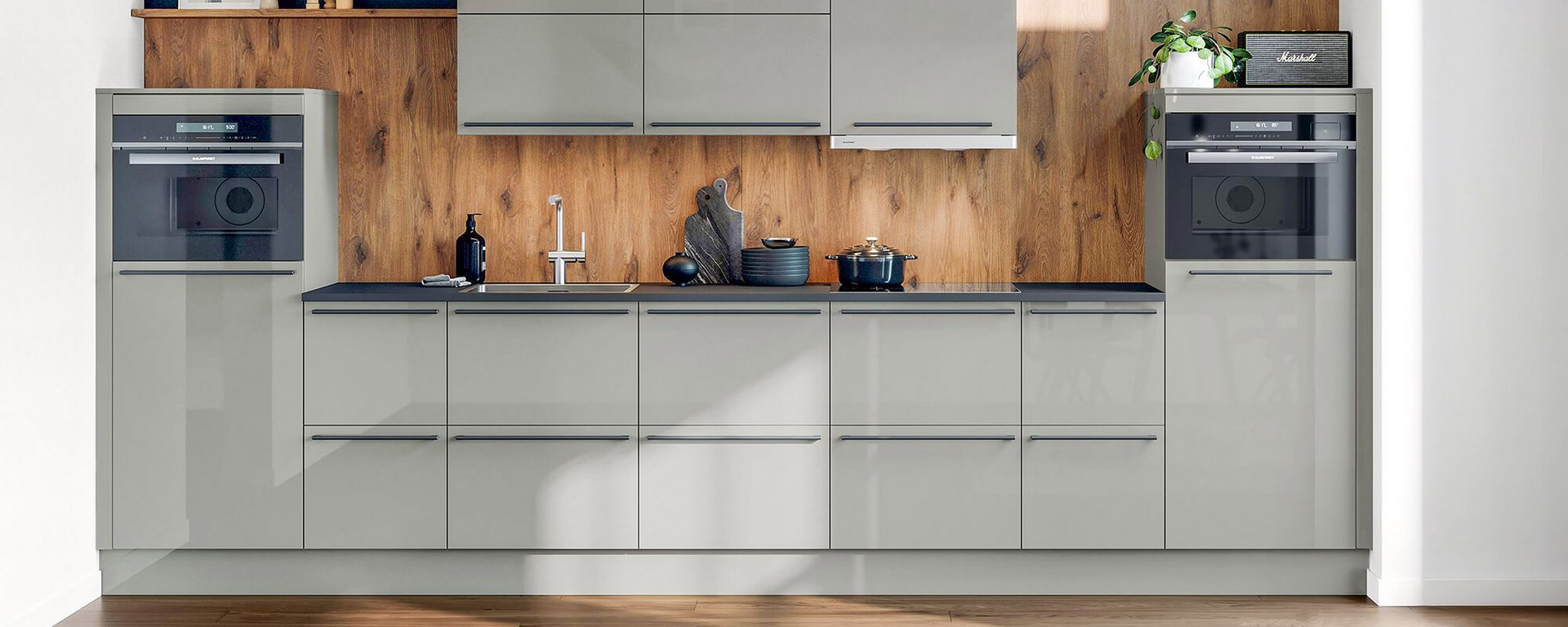 Küchenzeile mit glänzenden grauen Fronten, dunkler Arbeitsplatte, Rückwand in Holzoptik und zwei hoch eingebauten Elektrogeräten.