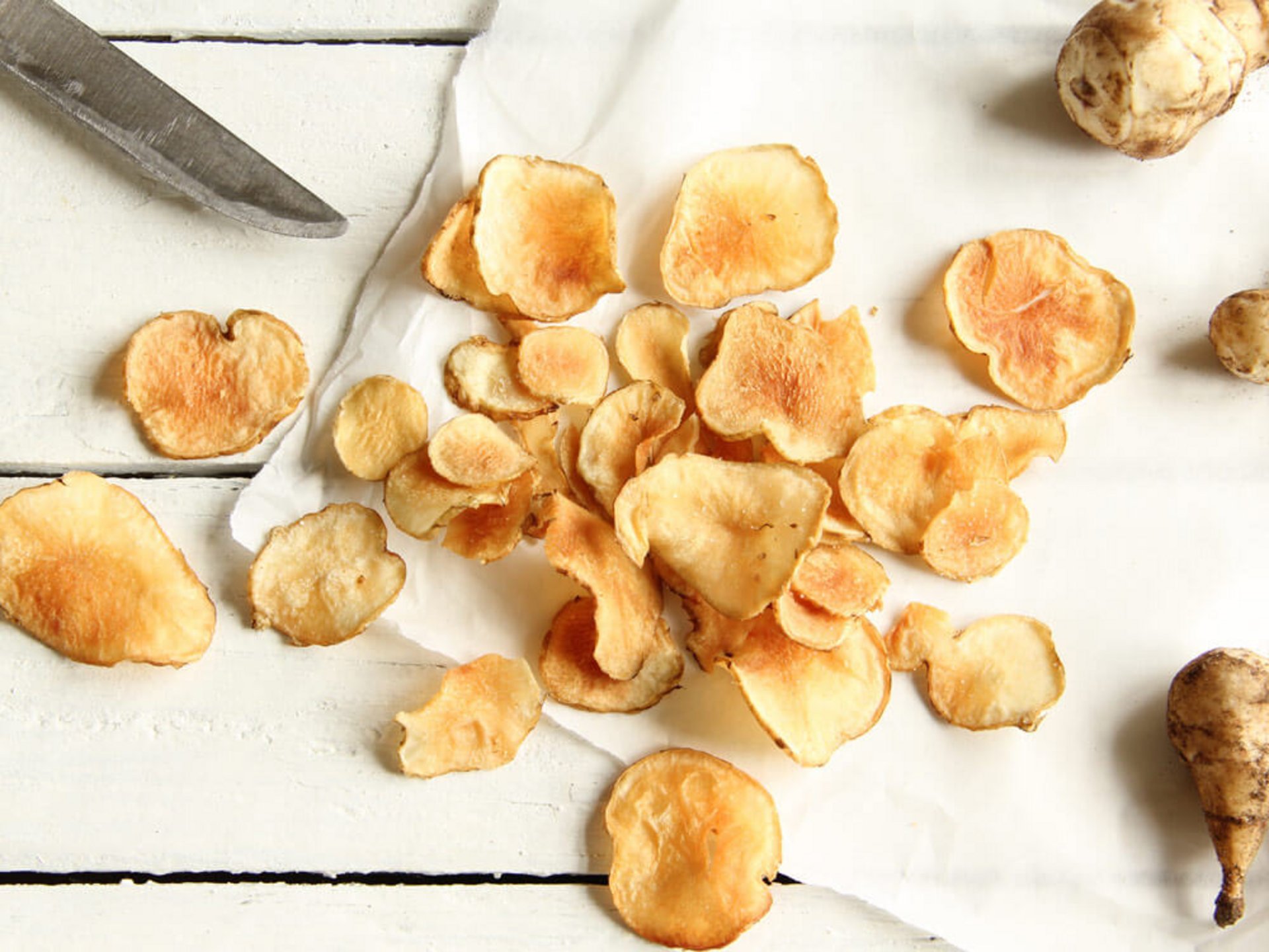 Hausgemachte, goldbraune Topinambur-Chips auf weißem Papier neben einem Messer – knuspriges Rezept mit Topinambur.