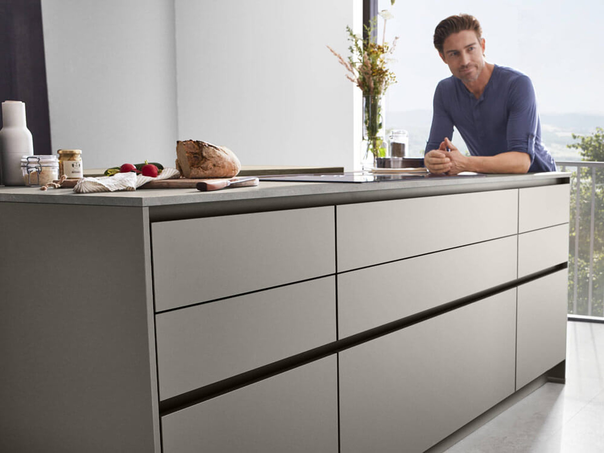 Moderner Mann lehnt an einer Kücheninsel mit pflegeleichten Anti-Fingerprint Fronten. Küchenplanung in Bayern.