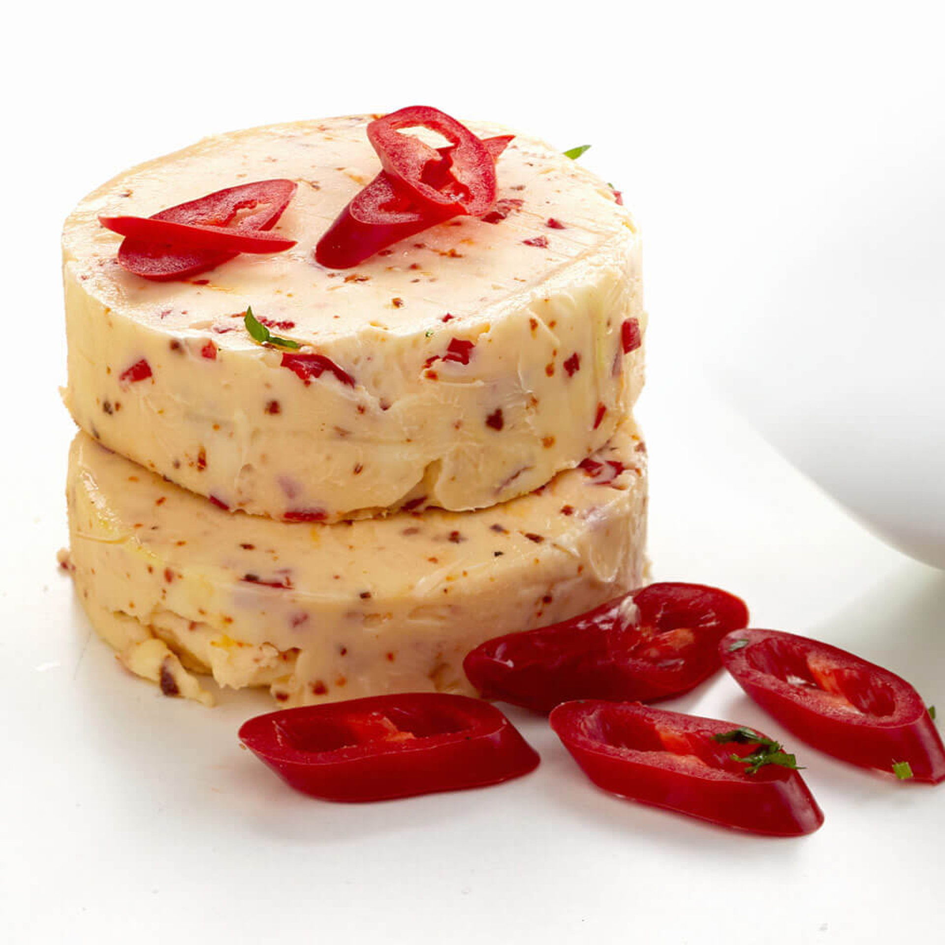 Zwei gestapelte Rollen würziger Chili-Butter, mit sichtbaren Chiliflocken, garniert mit frischen roten Chilischoten-Scheiben.