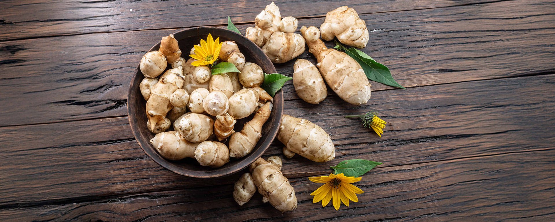 Topinambur-Knollen mit gelber Blüte in Holzschale auf dunklem Holztisch – Symbol für Ernte, Frische und Topinambur-Gesundheit.